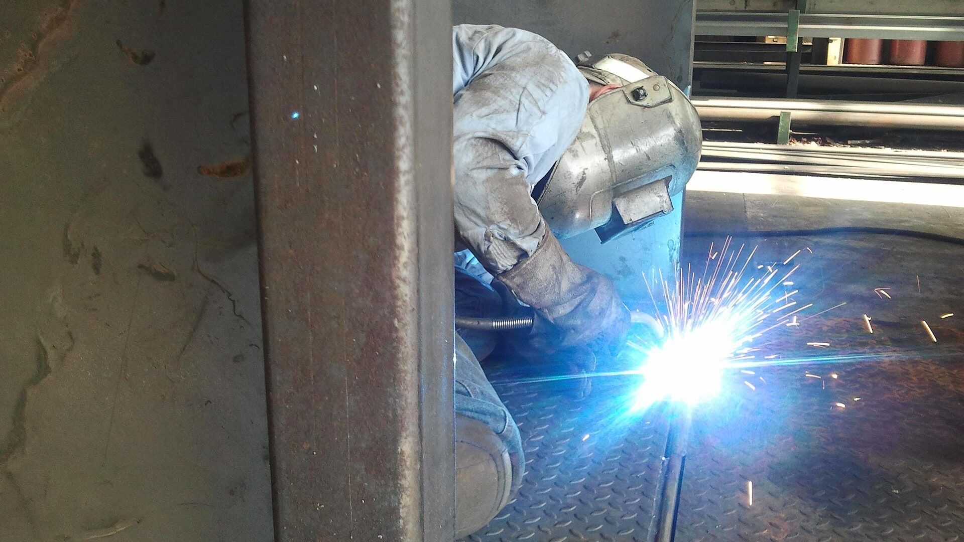 A welder crouched at the base of a steel column, arc lit, inside a fabrication bay