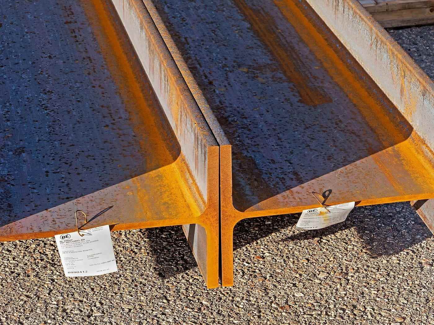 Rolled steel I-beams stacked on gravel in a stockyard, mill scale still orange
